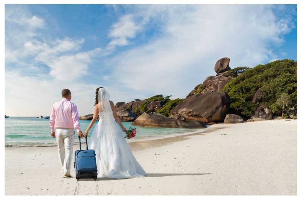Honeymoon couple walking along the beach with their suitcase.